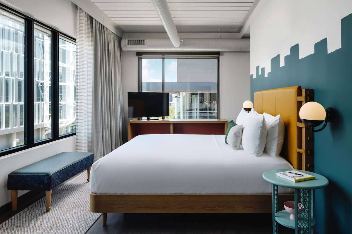Ein Hotelzimmer mit einem großen Bett, einer Bank, einem Fernseher, einem Nachttisch mit Lampe und Blick auf die Stadt durch das Fenster.