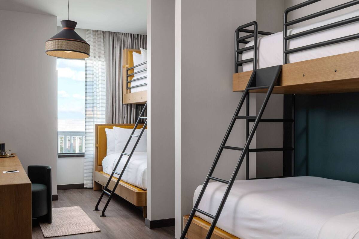 Ein kleines Hotelzimmer mit Stockbetten, einem Schreibtisch und einem großen Fenster mit Vorhängen.