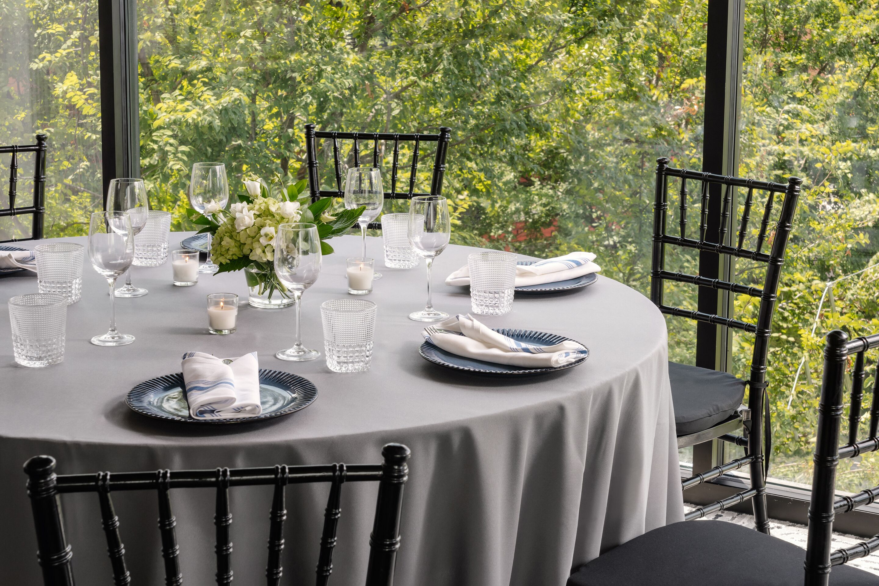 A round dining table with a white tablecloth, black chairs, and a view of greenery through large windows.