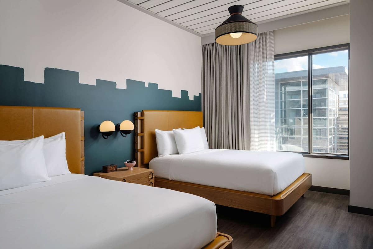Ein Hotelzimmer mit zwei Betten, einer Stadtlandschaftsmalerei, einem großen Fenster mit Vorhängen und einem modernen Deckenstil.