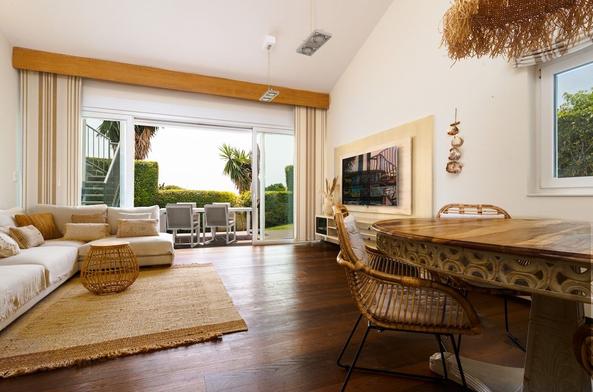 A modern living room with a sofa, dining table, and chairs, a large window with a view of greenery, and a ceiling light fixture.
