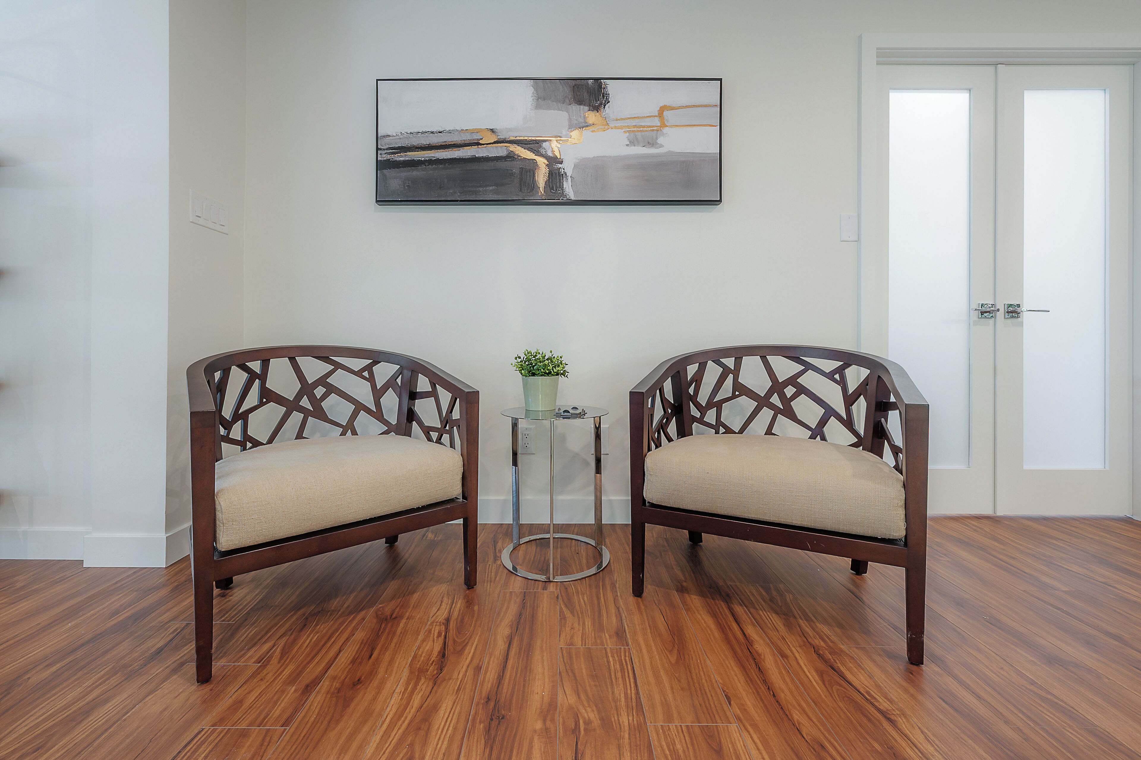 Ein Zimmer mit zwei Holzstühlen mit Kreuzgewebe-Lehnen, einem kleinen Tisch mit Glasplatte und einem Gemälde an der Wand.
