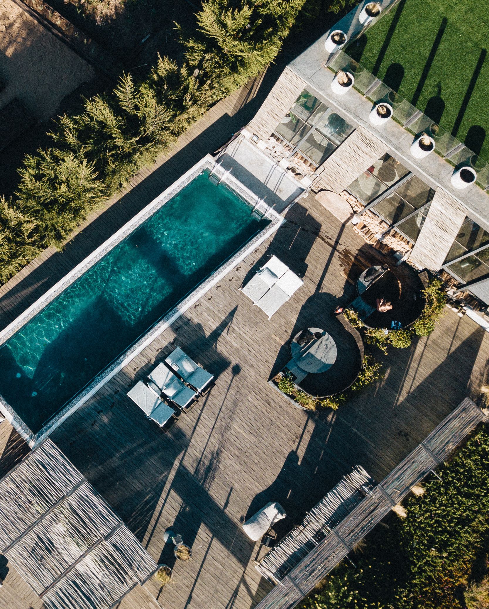 An aerial view of a rooftop pool area with clear water, surrounded by wooden decking, lounge chairs, and a circular seating area with a central fountain.