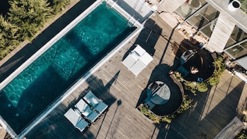 An aerial view of a rooftop pool area with clear water, surrounded by wooden decking, lounge chairs, and a circular seating area with a central fountain.