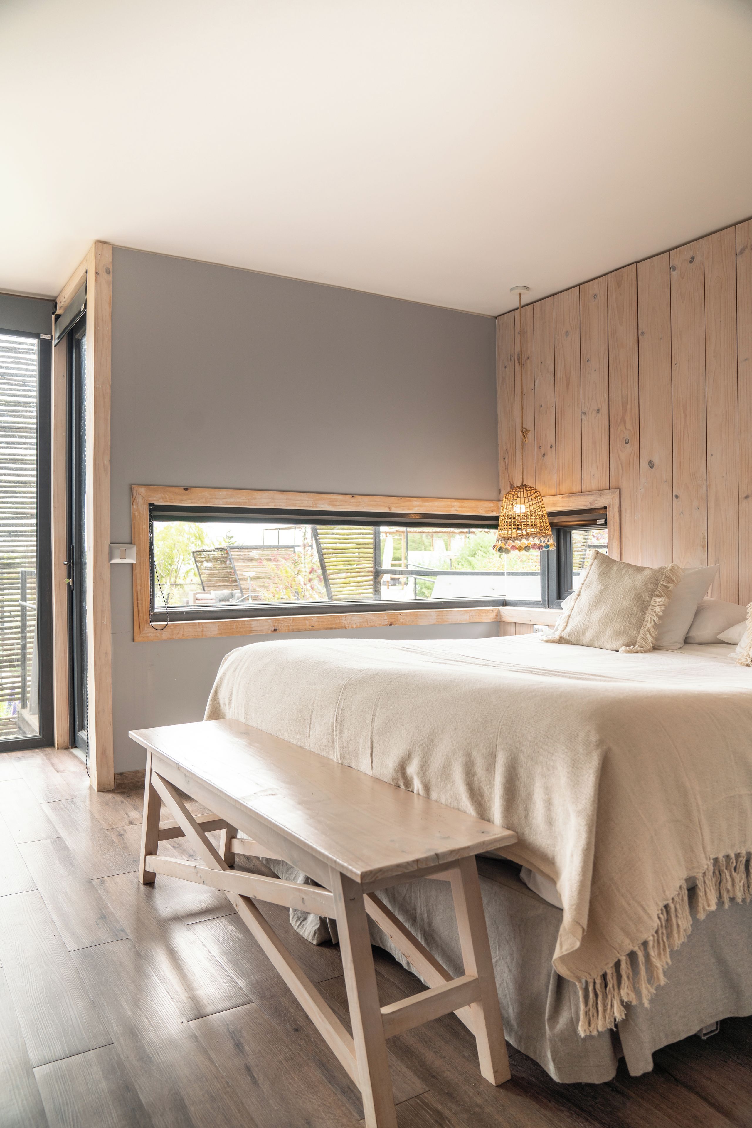 A bedroom with a large bed, a wooden bench, a window with a view, and a woven light fixture.