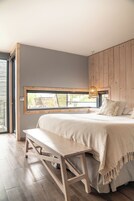 Un dormitorio con una cama grande, un banco de madera, un ventanal con vistas y una lámpara de techo tejida.