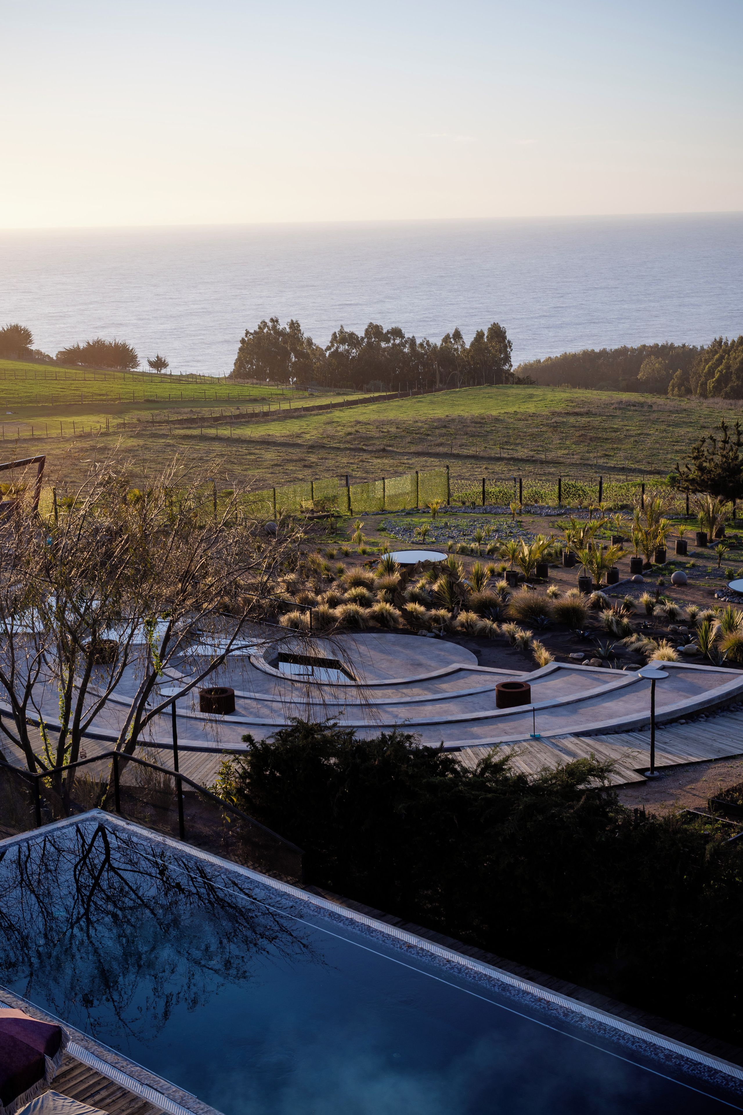 Zona de piscina con vista al océano, rodeada de jardines ajardinados y una valla.