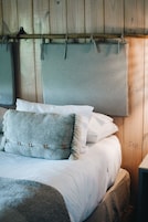 Una cama con sábanas blancas, un cojín de textura gris y un cabecero de madera.