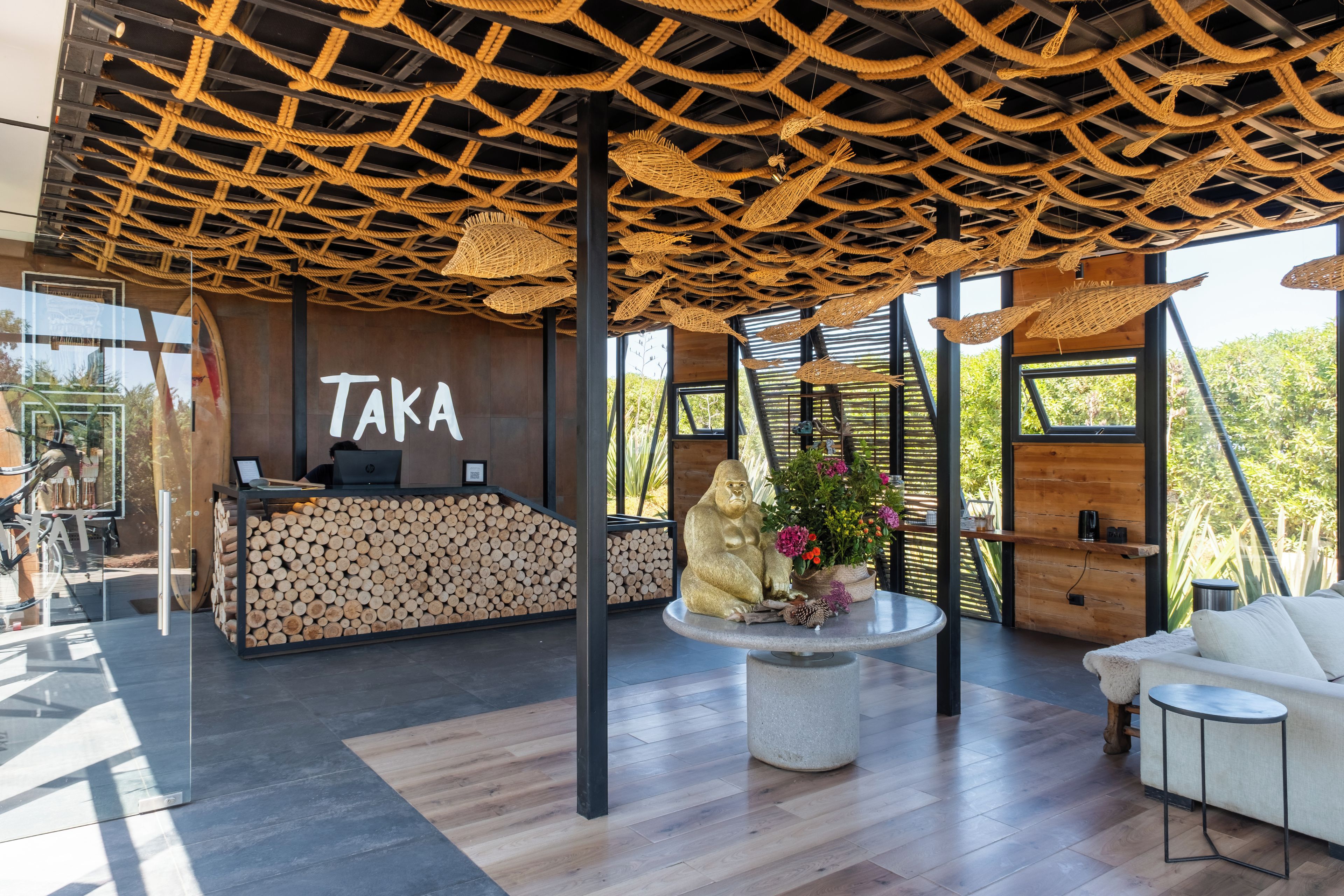 Zona de recepción con un letrero 'Taka', mostrador de recepción, una gran exhibición de un tronco de madera, un área de estar central con una mesa redonda y un sofá blanco.