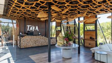 Zona de recepción con un letrero 'Taka', mostrador de recepción, una gran exhibición de un tronco de madera, un área de estar central con una mesa redonda y un sofá blanco.