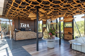 A reception area with a 'Taka' sign, a reception desk, a large wooden log display, a central seating area with a round table, and a white sofa. - Taka Matanzas (Navidad)
