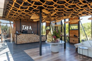 Zona de recepción con un letrero 'Taka', mostrador de recepción, una gran exhibición de un tronco de madera, un área de estar central con una mesa redonda y un sofá blanco.