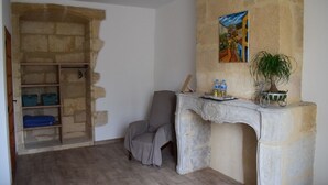 Una habitación con pared de piedra, un cuadro, una silla, una mesa con una planta y un estante con ropa doblada.