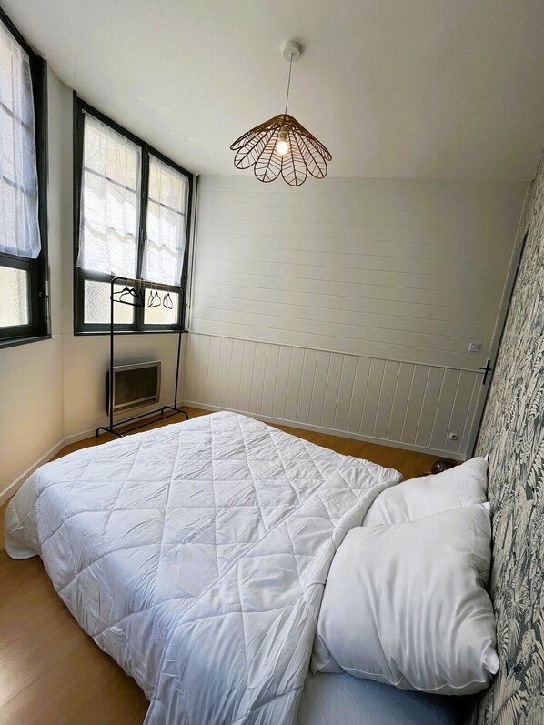 A bedroom with a bed, a hanging light fixture, a window with sheer curtains, and a radiator.