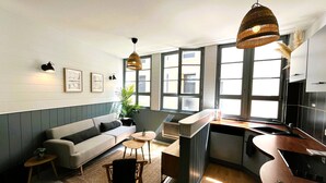 Una cocina moderna con estufa integrada, zona de comedor con sofá y una mesa pequeña, y ventanas amplias que dejan entrar luz natural.