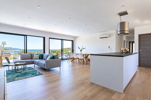 A modern interior with a kitchen island, dining area, and a sofa facing a large window overlooking a scenic view.