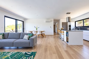 A modern living room with a grey sofa, a dining area with a white table and chairs, and a kitchen with a white countertop and bar stools.