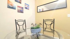 A dining area with a round glass table, two metal chairs, a potted plant, and a wall-mounted mirror. - Stay in this cozy and bright 2BR apartment, featuring a welcoming living room, modern décor, and plenty of natural light. Walk to Calle Ocho cafés, murals, and music, and reach Brickell in minutes. Perfect for families or groups exploring Miami (Miami)