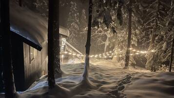 A snow-covered path leading to a cabin with a staircase and string lights.