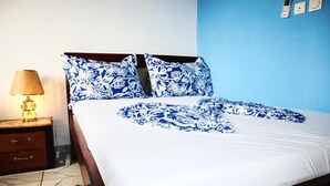 A bed with a white sheet and blue floral pillows, a wooden nightstand with a lamp, and a blue rug. - 1 Bed Studio Sleeps 2 - Canal Views, Balcony, Wifi (Douala)