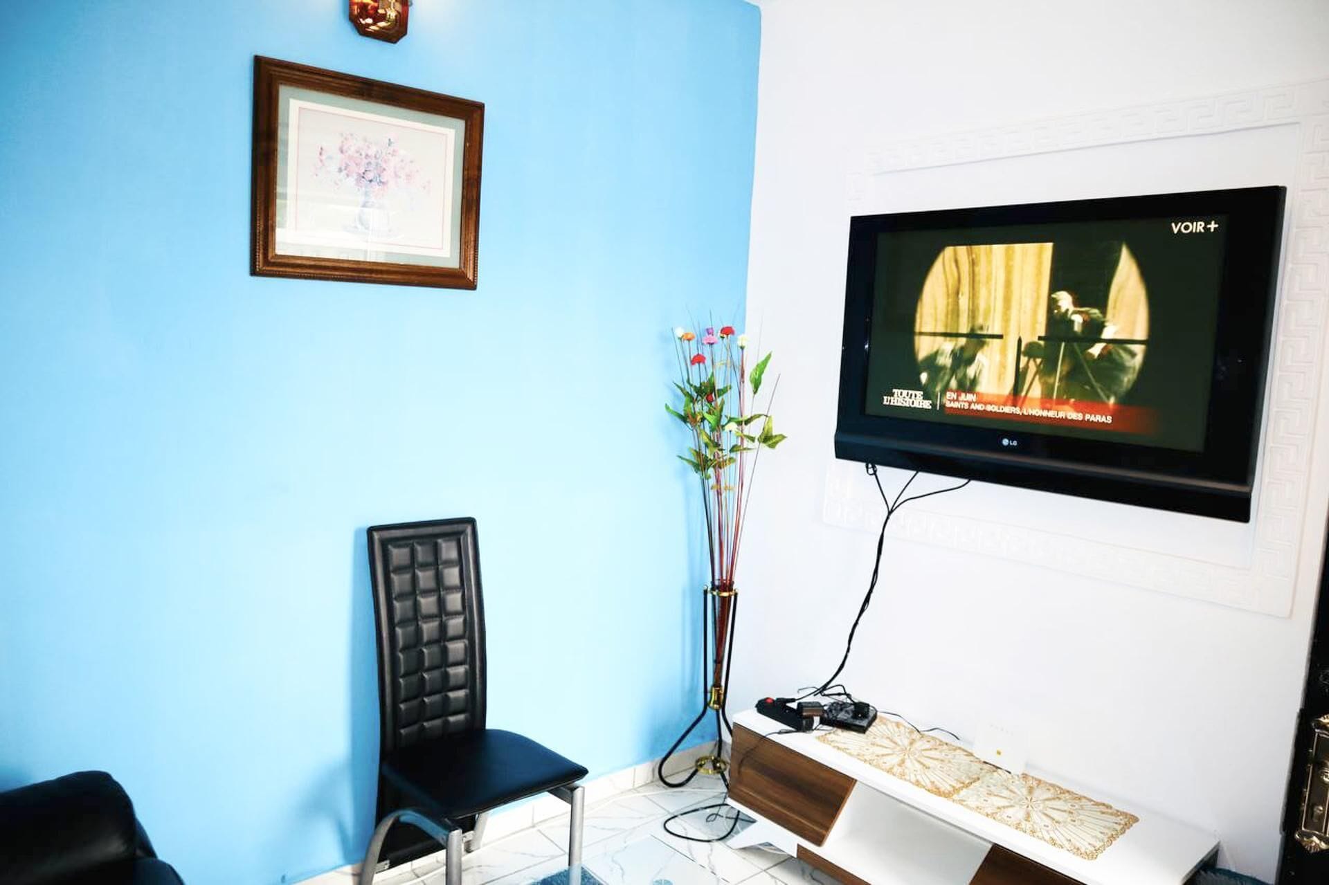 Une pièce avec un mur bleu, un tableau encadré, une chaise noire, une console blanche et un téléviseur fixé au mur.