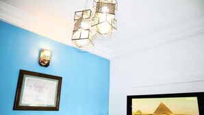A room with a blue wall, a framed picture, a wall-mounted light fixture, a TV displaying a desert scene, and a plant with flowers. - 1 Bed Studio Sleeps 2 - Canal Views, Balcony, Wifi (Douala)
