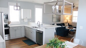 A modern kitchen with white cabinets, a built-in oven, and a dishwasher. There is a dining area with a wooden table and a potted plant.