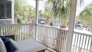 A balcony with a white cushioned bench, white railings, and a view of palm trees and a pool area.
