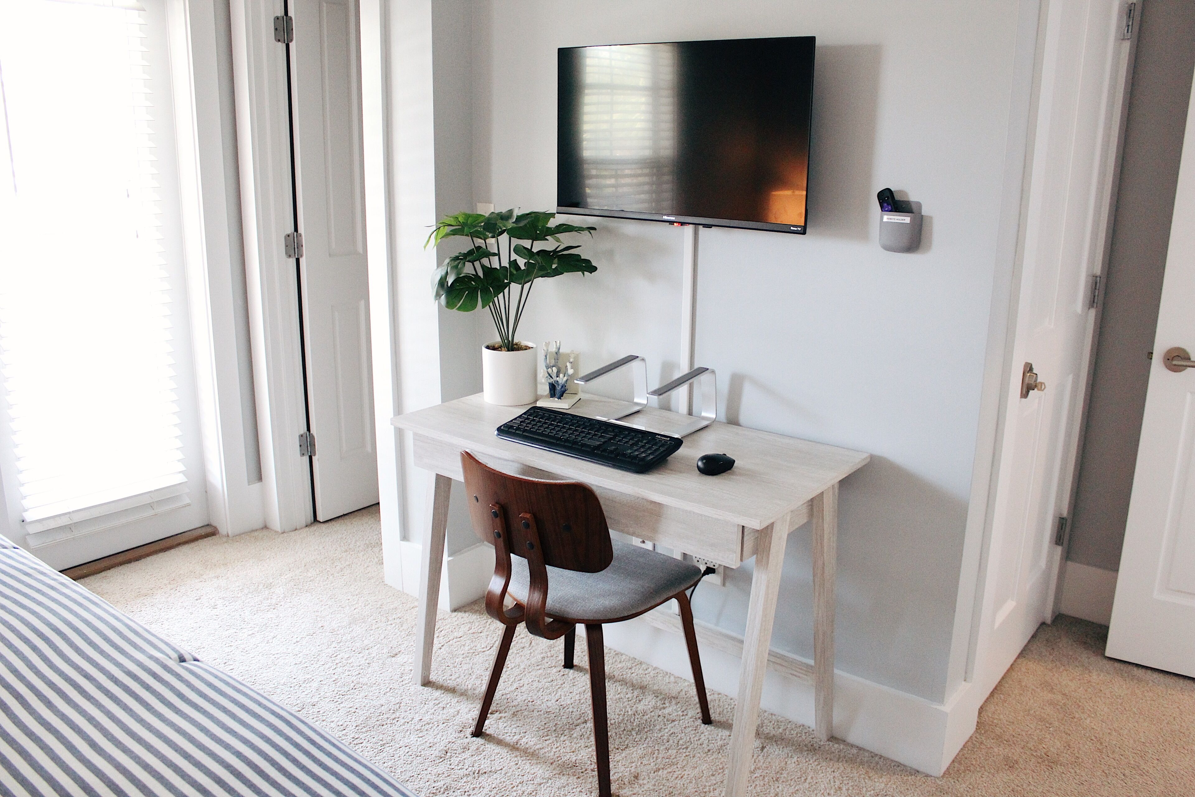 Un dormitorio con cama, escritorio, computadora, silla, planta y televisor en la pared.