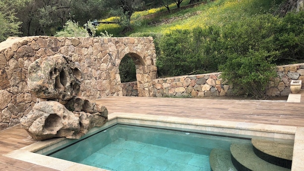 A rectangular pool with a stone wall and archway, surrounded by a wooden deck and greenery. - Caseddu in the shade of the Olive Trees, just a few steps from the sea. (Olmeto)