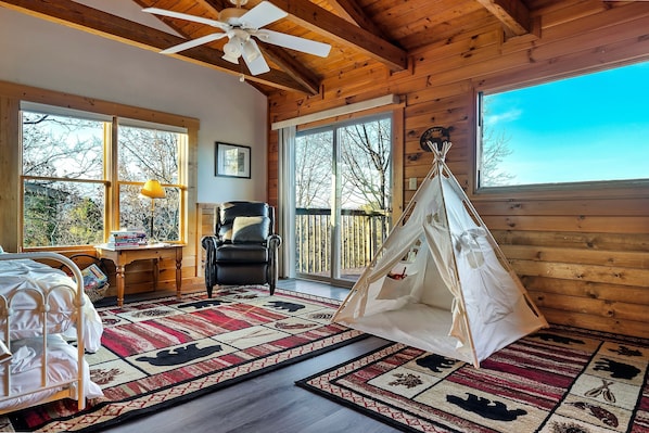 A cozy room with a bed, a desk, a chair, a hanging chair, and large windows with a view of trees.
