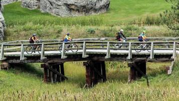 Un ponte di legno che sovrasta un prato, con dei ciclisti che lo attraversano.