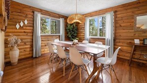 Interior de una cabaña de madera con una mesa de comedor, sillas, una lámpara colgante y ventanas amplias.