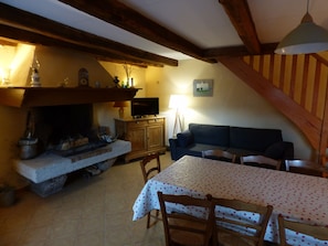A cozy living room with a fireplace, wooden beams, a sofa, a dining table with chairs, and a television. - Gîte chez Sylvie - La Brousse - Pied du Mont Lozère (FRAISSINET DE LOZERE)