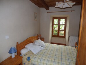 A bedroom with a wooden bed, a nightstand with a lamp, a window with a view of greenery, and a radiator. - Gîte chez Sylvie - La Brousse - Pied du Mont Lozère (FRAISSINET DE LOZERE)