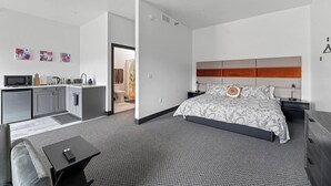 A modern hotel room with a bed, bedside tables, a desk, a kitchenette, and a bathroom. - Downtown Executive Apartment - Pyrite (Spearfish)