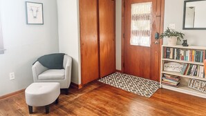 Um quarto com porta de madeira, uma cadeira branca, um pequeno banquinho, uma estante com livros e um tapete estampado.
