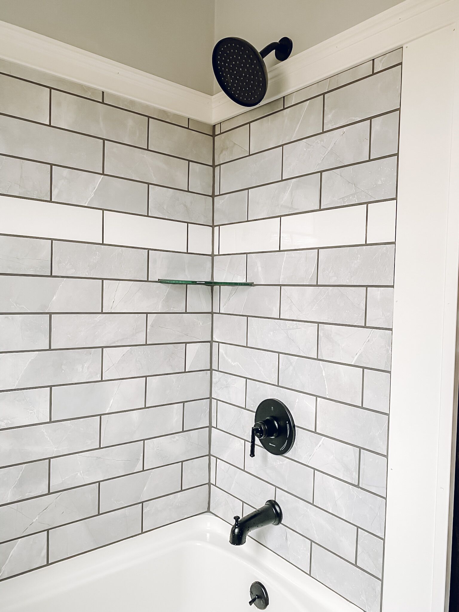 Baño en esquina con azulejos de mármol blanco, cabezal de ducha, grifo y repisa de vidrio.