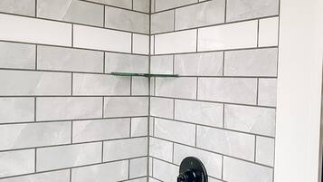 Baño en esquina con azulejos de mármol blanco, cabezal de ducha, grifo y repisa de vidrio.
