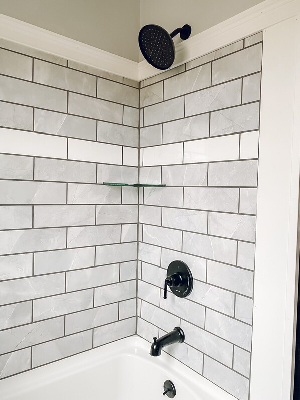 A corner bathroom with white marble tiles, a showerhead, a faucet, and a glass shelf.
