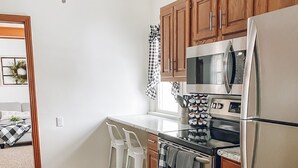 Cozinha moderna com armários de madeira, geladeira de aço inoxidável, micro-ondas e forno. Há dois banquetas brancas e um tapete preto no chão.