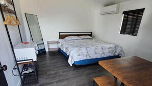 Quarto compacto com cama, uma pequena escrivaninha, espelho e janela com persianas.