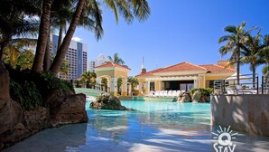 A hotel pool area with clear blue water, palm trees, and a view of a city skyline. - Chevron – 3 Bedroom Superior Unit With Ocean Views (Surfers Paradise)