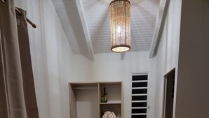 A small, well-lit room with a wooden floor, a wardrobe with open doors, a hanging light fixture, and a bed with a patterned cover. - Building (Les Anses-d'Arlet)