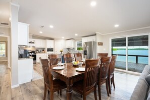 A modern kitchen with white cabinets, a stainless steel refrigerator, and a dining area with wooden chairs and a large table. - Angler`s Cove · Waterfront House with Amazing Views & Giant Deck (Hemphill)