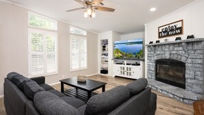 Una sala de estar con chimenea, ventilador de techo y televisor sobre un mueble blanco.