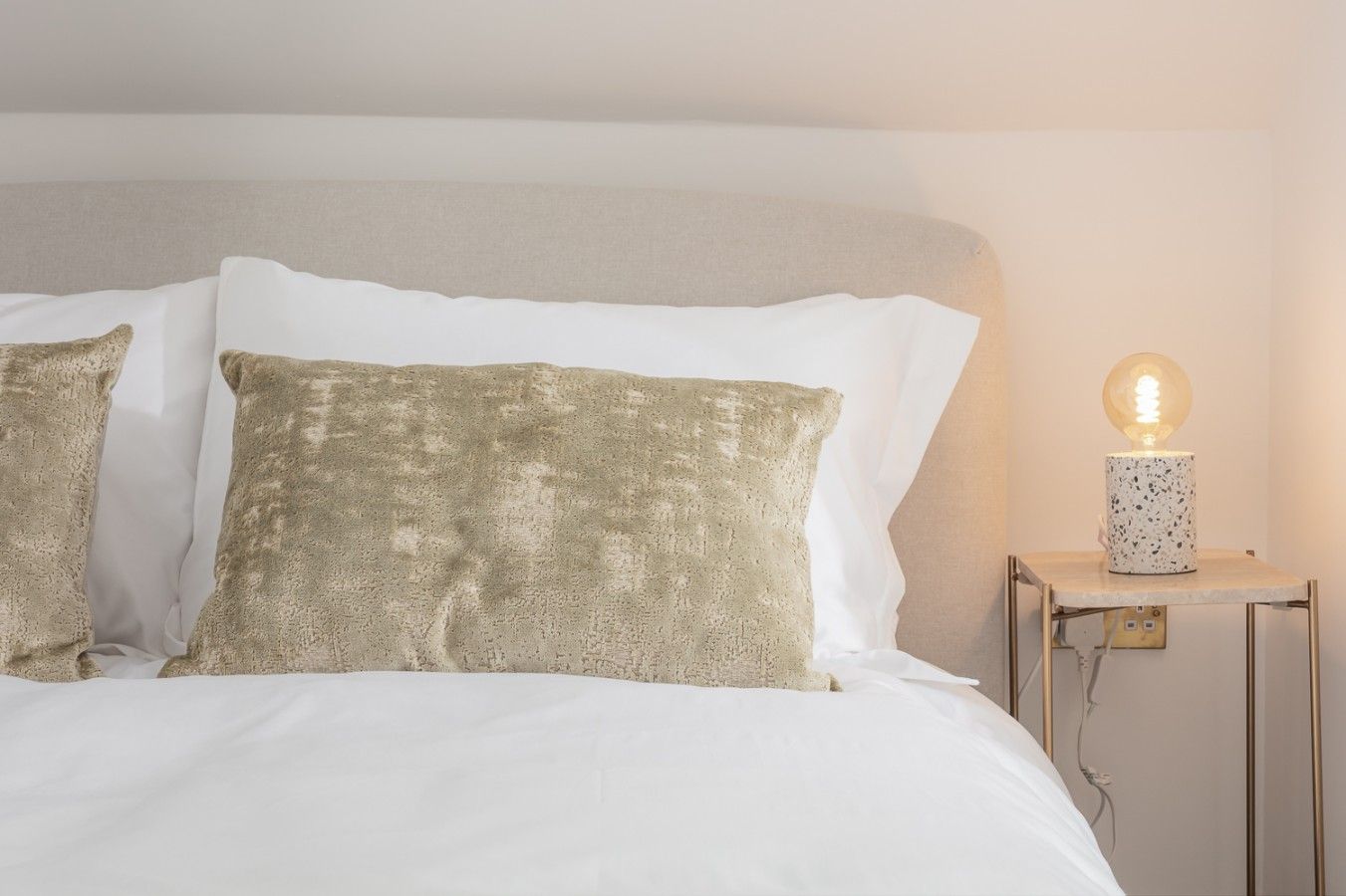 Ein Bett mit weißer Bettwäsche und zwei strukturierten Kissen, ein Nachttisch mit einer Lampe sowie ein Lichtschalter an der Wand.