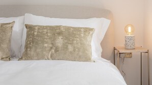 Un lit avec une literie blanche et deux coussins à texture, une table de chevet avec une lampe et un interrupteur mural.