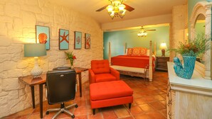 A room with a stone wall, a bed with red bedding, a desk with a lamp, a chair, and a potted plant.