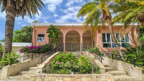 A pink building with a stone staircase leading to an arched entrance, surrounded by tropical plants and palm trees.